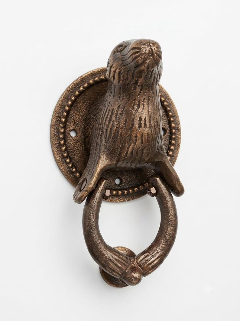May include: A bronze-colored metal door knocker featuring a sculpted seal head mounted on a circular backplate with a beaded edge. The seal's front flippers extend downward to hold a circular ring that serves as the striking part of the mechanism.