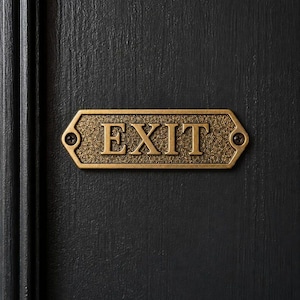 May include: A rectangular brass-toned metal exit sign with a textured background and raised lettering reading EXIT. The sign features beveled edges and is secured to a dark, vertically grained surface by two visible screws on the left and right sides.