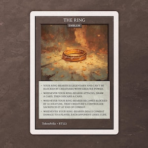 Puede incluir: Una carta con el emblema "THE RING", que muestra una ilustración de un anillo dorado. El texto describe las habilidades del anillo, como robar cartas y causar daño de combate. El fondo es borroso y artístico.