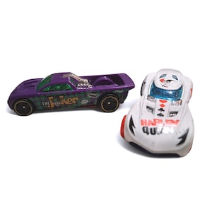 Può includere: Due macchinine: un'auto viola con grafica "The Joker" e un'auto bianca con grafica "Harley Quinn". L'auto viola ha un parabrezza verde e ruote nere. L'auto bianca ha dettagli rossi e finestrini blu.