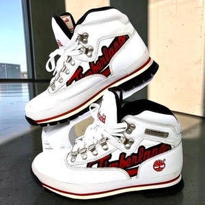 May include: A pair of white high-top boots with black interior lining and red accents. The side features the word Timberland in red script lettering with a red tree logo. The boots have silver metal eyelets, white laces, and a thick black sole.