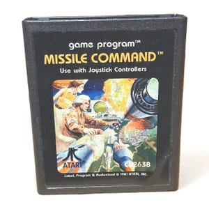 Puede incluir: Cartucho de juego Atari Missile Command negro. La etiqueta presenta el título del juego en amarillo y un gráfico de lanzamiento de misiles. También es visible el texto "Use with Joystick Controllers". El logotipo de Atari está en la parte inferior.