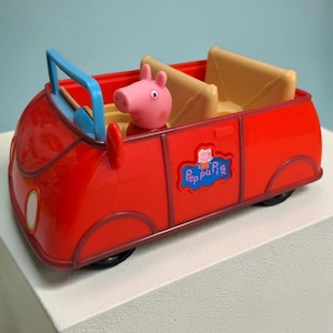 May include: A bright red plastic toy car featuring a Peppa Pig character figure seated inside. The vehicle has a light blue windscreen frame, tan interior seats, and a blue logo on the side panel with the text Peppa Pig in white letters.