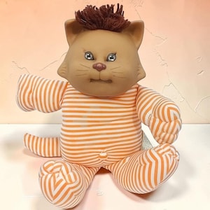 May include: A plush cat doll with a tan face, brown yarn hair, and embroidered facial features. The body is covered in an orange and white horizontal striped fabric, featuring a small round belly button detail and a matching striped tail.