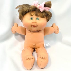Pode incluir: Uma boneca com corpo de tecido macio em tom de pêssego e cabeça de vinil. A boneca tem cabelo castanho com um laço de tecido rosa claro, olhos verdes e uma pequena covinha na bochecha esquerda. A boneca tem uma expressão de boca aberta.