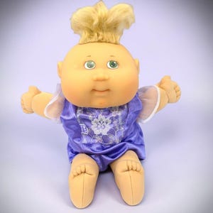 May include: A doll with a round, peach-toned face, green eyes, and blonde hair styled in a high tuft. The doll wears a purple satin-like outfit featuring a white floral lace panel on the chest, sheer white sleeves, and a small tag labeled CPK.