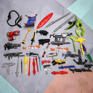 Könnte beinhalten: Eine Sammlung verschiedener Miniatur-Spielzeugwaffen und Zubehörteile, darunter diverse Blaster, Schwerter, Speere und futuristische Ausrüstung in den Farben Silber, Schwarz, Rot, Gelb, Grün und Blau, angeordnet auf einer flachen Oberfläche.