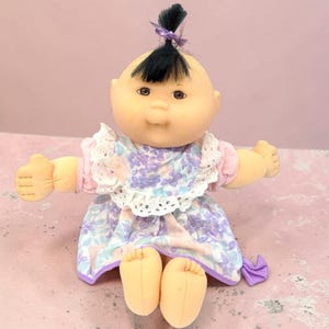 May include: A soft-sculpture doll with a round face, brown eyes, and a single tuft of black hair tied with a purple ribbon. The doll wears a dress featuring a floral pattern in shades of purple, blue, and white, accented with a white eyelet lace collar.