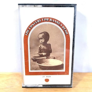 May include: A cassette tape for "The Concert for Bangla Desh" album. The cover features a sepia-toned image of a child with a bowl. The text is in a red and white arched design. The Apple logo is at the bottom.