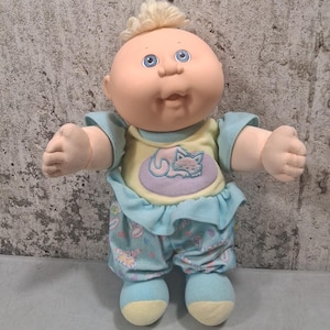 Könnte beinhalten: Eine Vintage-Cabbage-Patch-Kid-Puppe mit rundem Gesicht, blauen Augen und einem Büschel blondem Haar. Die Puppe trägt ein hellblaues und gelbes Outfit mit einer Katzenzeichnung auf der Vorderseite. Die Arme der Puppe sind ausgestreckt.