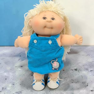 Peut inclure: Une poupée à la peau claire, aux joues roses et aux cheveux en laine blonde texturée. La poupée porte une salopette en velours côtelé bleu vif avec deux boutons blancs sur la poitrine et un petit éléphant brodé, ainsi que des baskets blanches à rayures bleues.