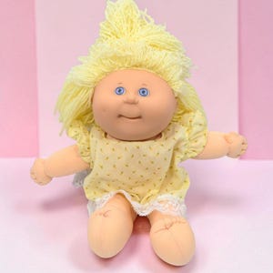 Peut inclure: Une poupée en tissu avec une peau claire, des yeux bleus et des cheveux en laine jaune vif. La poupée porte un haut sans manches jaune pâle avec un petit motif floral, une bordure en dentelle blanche à l'ourlet et des manches à volants.