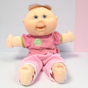 Pode incluir: Uma boneca Cabbage Patch Kids com rosto redondo, pele clara e cabelo castanho. A boneca veste uma camisa rosa de manga curta com bolinhas brancas e um emblema verde com o logótipo CPK, combinada com calças rosa claro. Tem olhos verdes e um sorriso.