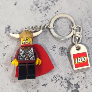 May include: A LEGO keychain featuring a Viking minifigure with a gold horned helmet, a yellow face with a beard, a silver and black patterned torso, and a red cape. The keychain includes a silver metal ring and a white tag with the red LEGO logo.