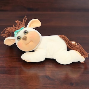 May include: A plush doll featuring a cream-colored body with brown yarn hair and a matching brown yarn tail. The doll has a soft, rounded face with green eyes, a small nose, and pink cheeks, accented by a small teal bow in its hair.