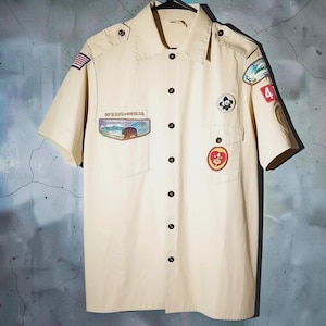 May include: A tan short-sleeved button-down shirt featuring a collar, shoulder epaulets, and two chest pockets. The shirt displays various embroidered patches, including a Boy Scouts of America emblem and a United States flag on the sleeve.