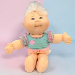 Könnte beinhalten: Eine Puppe mit heller Haut, blondem Haar und braunen Augen. Die Puppe trägt ein türkisfarbenes Oberteil mit einem rosafarbenen, blumenförmigen Aufnäher mit den Buchstaben CPK, akzentuiert durch gerüschte Blumenärmel und einen rosafarbenen Saum.