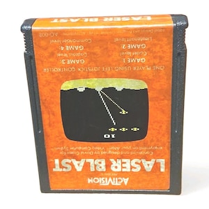 Puede incluir: Un cartucho de videojuego Laser Blast vintage de Activision. La etiqueta naranja presenta un gráfico de pantalla de juego con líneas blancas y formas amarillas. El cartucho tiene el título del juego y el logotipo de la empresa, con los niveles del juego.