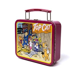 Peut inclure: Une boîte à déjeuner rectangulaire en métal violet avec une illustration colorée des personnages du dessin animé Top Cat dans une ruelle. Le motif inclut un policier, plusieurs chats et le texte Top Cat imprimé en blanc sur un ciel jaune.