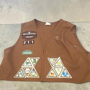 May include: A brown Girl Scouts USA vest from Vermont, featuring the number 211, a green arch patch, and several triangular embroidered badges on the front panels. A small gold-toned pin is attached to the upper right side of the garment.