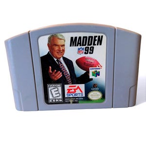 May include: A gray Nintendo 64 game cartridge for Madden NFL 99. The label features a portrait of a man in a suit, a football, and the NFL and Nintendo logos. The game is rated "Everyone" by the ESRB.