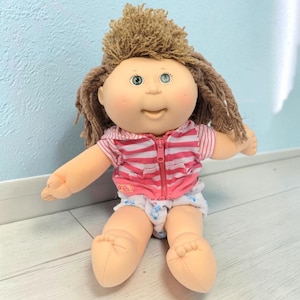 May include: A doll with light skin, green eyes, and brown yarn hair styled in a messy top knot. The doll wears a pink and white horizontal striped zip-up hoodie with short sleeves and white shorts featuring a small blue pattern of figures.