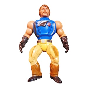 Könnte beinhalten: Vintage Actionfigur eines muskulösen Mannes mit braunen Haaren und Schnurrbart. Die Figur trägt ein blau-gelbes Outfit mit braunen Stiefeln und Handschuhen. Das Spielzeug hat eine Retro-Ästhetik.