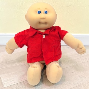May include: A doll with a round, light-toned fabric head, bright blue eyes, and a small nose. The doll has a soft, stuffed body and is wearing a short-sleeved, button-down red shirt with a single white button visible at the center of the chest.