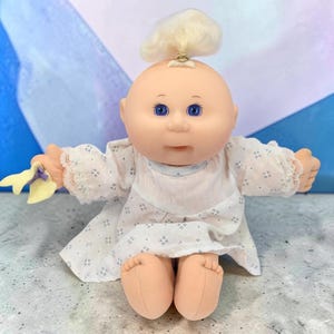 Pode incluir: Uma boneca com cabeça redonda, pele clara e olhos azuis. Tem cabelo loiro preso num coque com uma fita dourada. A boneca veste um vestido branco de manga comprida com um pequeno padrão geométrico azul e renda nos punhos. Segura um acessório de tecido amarelo na mão esquerda.