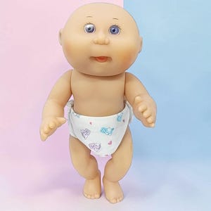 Peut inclure: Une poupée bébé chauve avec une tête ronde, un teint clair et des yeux bleus. La poupée a un petit nez, une bouche légèrement ouverte et des joues roses. Elle porte une couche blanche avec un motif répétitif de petits blocs bleus et roses.