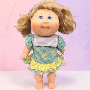 Puede incluir: Una muñeca con cabello rubio peinado en dos coletas, ojos azules y mejillas sonrosadas. La muñeca viste un vestido con un corpiño verde azulado, un cuello morado claro y una falda amarilla, decorados con un patrón colorido de soles y corazones.