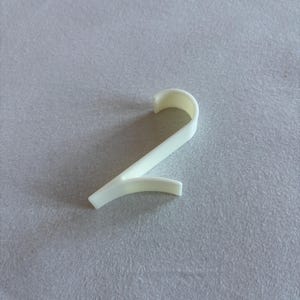 Puede incluir: Objeto blanco de plástico con forma de número dos. El objeto tiene una parte superior curva y una inferior recta, con una ligera curva en el extremo. Podría ser un artículo decorativo o un suministro artesanal.