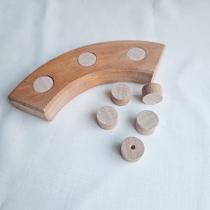 Könnte beinhalten: Ein gebogener Holzblock mit drei kreisförmigen Vertiefungen und fünf separaten kleinen Holzzylindern, von denen einer ein kleines Loch in der Mitte aufweist. Das Holz hat eine natürliche hellbraune Maserung und ein glattes, mattes Finish.