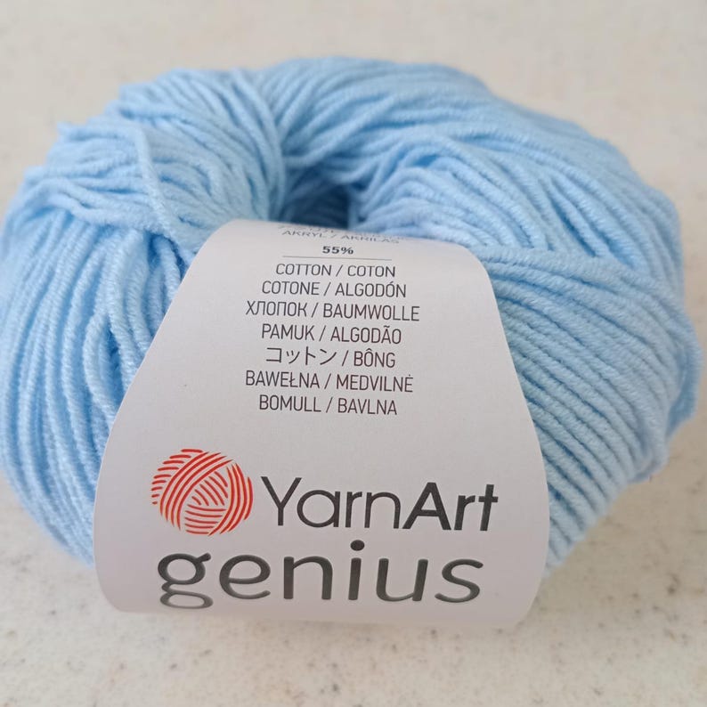 Pode incluir: Um novelo de fio azul claro com uma etiqueta de papel branco apresentando o nome da marca YarnArt genius em texto preto e um log&oacute;tipo de novelo vermelho. A etiqueta indica 55% algod&atilde;o em v&aacute;rios idiomas, detalhando a composi&ccedil;&atilde;o do fio.