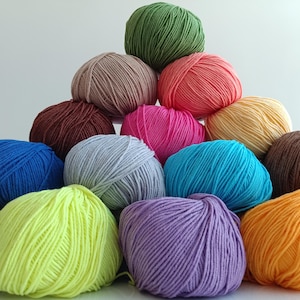 Pode incluir: Uma pilha piramidal de doze novelos de l&atilde; em v&aacute;rias cores s&oacute;lidas, incluindo verde-lim&atilde;o, coral, magenta, cinza-claro, azul brilhante, roxo, laranja, marrom, bege, marrom escuro e verde-oliva, dispostos em um grupo denso e multicamadas.