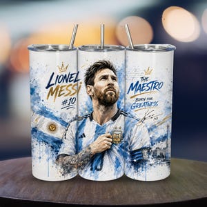 Peut inclure: Un gobelet cylindrique en acier inoxydable avec une paille en métal, orné d'un portrait de Lionel Messi portant le maillot argentin. Le design présente des éclaboussures bleu et blanc, avec les inscriptions Lionel Messi #10, The Maestro, Born for Greatness.