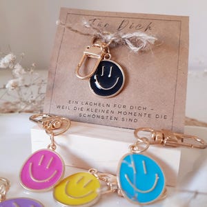 Könnte beinhalten: Eine Sammlung von goldfarbenen Metall-Schlüsselanhängern mit runden, bunten Emaille-Smiley-Anhängern in Pink, Gelb, Blau, Lila und Schwarz. Die Anhänger sind an einer braunen Karte mit dem Text Für Dich und einem deutschen Satz befestigt.