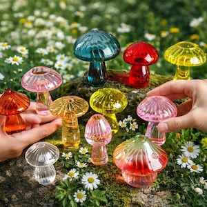 Może przedstawiać: Kolekcja kolorowych szklanych figurek grzybów w różnych rozmiarach i odcieniach, w tym czerwonym, żółtym, różowym i turkusowym. Grzyby są prezentowane na mszystej powierzchni z białymi kwiatami w tle.
