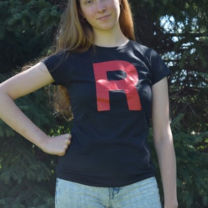 Può includere: Maglietta nera con una grande "R" rossa. La persona indossa pantaloncini di jeans blu chiaro. Lo sfondo presenta un fogliame verde.