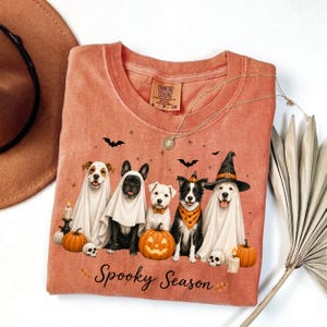 May include: A rust-colored t-shirt featuring a graphic of five dogs, some wearing ghost costumes and witch hats, surrounded by pumpkins, skulls, and bats. The text "Spooky Season" is printed below the dogs. The shirt has a Comfort Colors label.