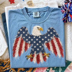 May include: A light blue Comfort Colors t-shirt featuring an embroidered American flag eagle design. The eagle's wings display the stars and stripes of the U.S. flag. The shirt is folded, with other shirts visible in the background.