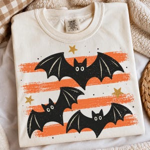 May include: A cream-colored short-sleeve t-shirt featuring a graphic print of three black bats flying over horizontal orange brushstroke stripes. The design includes small gold stars and black dots scattered across the fabric. The shirt has a Comfort Colors label.