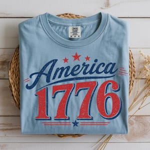 Pode incluir: Camiseta azul claro com a palavra "America" em escrita azul marinho e o número "1776" em vermelho, com estrelas e listras vermelhas. A camiseta está dobrada sobre uma cesta de vime.