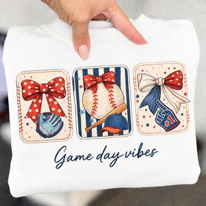 May include: White sweatshirt with three baseball-themed graphics. Each graphic features a red, white, and blue bow, baseball elements, and stars. The text "Game day vibes" is written in cursive below the graphics.