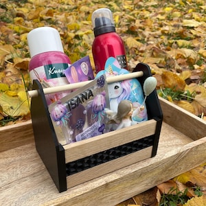 Könnte beinhalten: Ein Holzkasten mit schwarzem Griff und hellem Holzsockel, gefüllt mit Schönheitsprodukten. Der Kasten steht auf einem Holztablett, umgeben von Herbstlaub. Produkte umfassen eine rosa Flasche, eine rote Flasche und eine Einhorn-Karte.