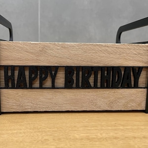 May include: A wooden box with black handles and trim, featuring the words "HAPPY BIRTHDAY" cut out in black across the front. The box has a light brown, wood-grain appearance.