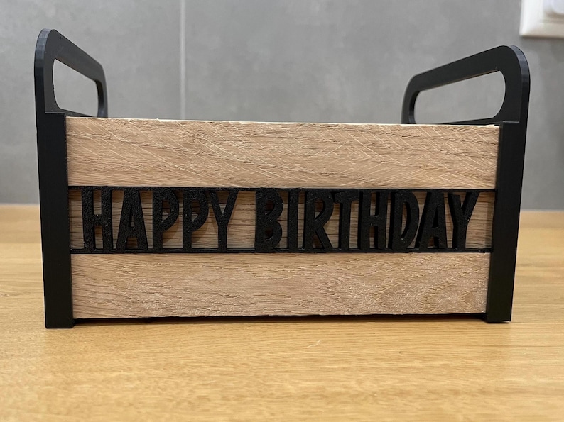 May include: A wooden box with black handles and trim, featuring the words "HAPPY BIRTHDAY" cut out in black across the front. The box has a light brown, wood-grain appearance.