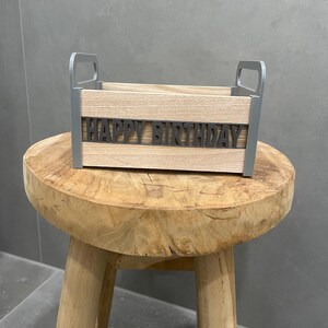 May include: A wooden box with gray handles and the words "HAPPY BIRTHDAY" in gray letters. The box is made of light-colored wood and sits on a small, round wooden stool.