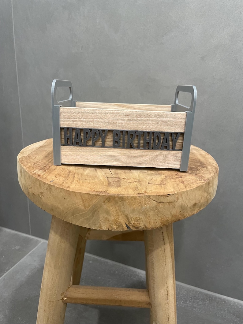 May include: A wooden box with gray handles and the words "HAPPY BIRTHDAY" in gray letters. The box is made of light-colored wood and sits on a small, round wooden stool.