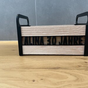 May include: A wooden box with black handles and trim. The front of the box features the text "ALINA 30 JAHRE" in black, cut-out letters. The box has a light wood grain finish and is sitting on a wooden surface.